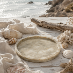 Luxury round beige marble tray styled on a beach with seashells, driftwood, and soft coastal decor.
