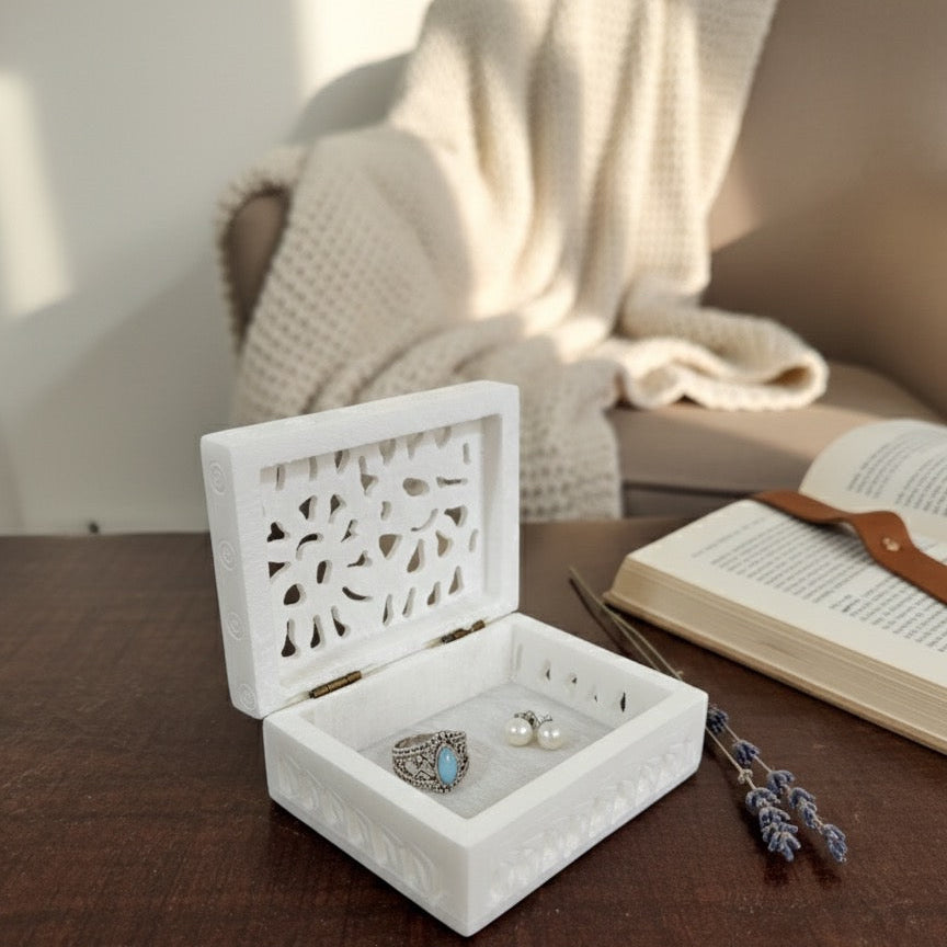 Marble jewelry box with a decorative lid, open on a dark wooden table. Inside the box are a silver ring with a blue stone and a pair of pearl stud earrings.